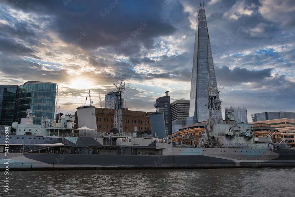 HMS Belfast, a historic Royal Navy warship, moored on the River Thames in London, with The Shard and modern office buildings under a partly cloudy sky.