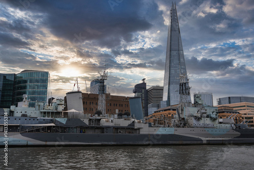 Obraz na płótnie HMS Belfast, a historic Royal Navy warship, moored on the River Thames in London, with The Shard and modern office buildings under a partly cloudy sky. w salonie