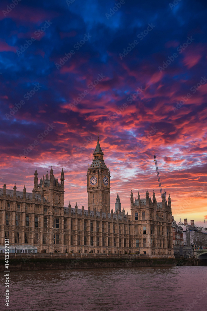 Elizabeth Tower, part of the Palace of Westminster, illuminated by a vibrant sunset. The River Thames reflects the colorful sky, with a crane visible nearby.