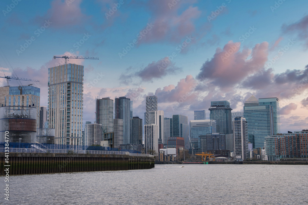 Modern skyscrapers of Canary Wharf in London with the River Thames in the foreground, construction cranes, and a partly cloudy early evening sky.