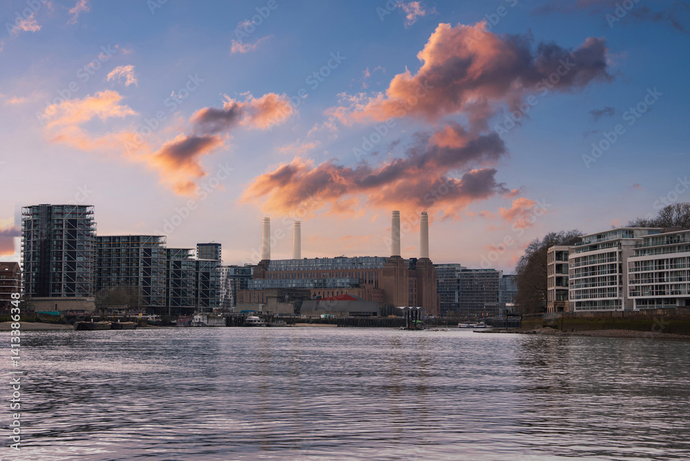 Battersea Power Station with its four chimneys stands amid modern buildings. The River Thames flows in the foreground under a vibrant sunset sky.