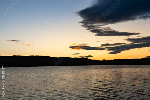 A calm lake reflects the rich colors of sunset as the blue hour envelops the landscape. Dark silhouettes of mountains rise against the vibrant sky in Dalarna, Sweden