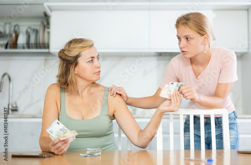 Murais de parede Focused woman received salary counting money at kitchen table while teenage daughter patiently waiting for pocket money