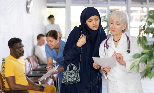 Experienced aged female doctor showing results of medical examination and explaining prescribed treatment to young asian woman wearing traditional muslim dress in lobby of clinic