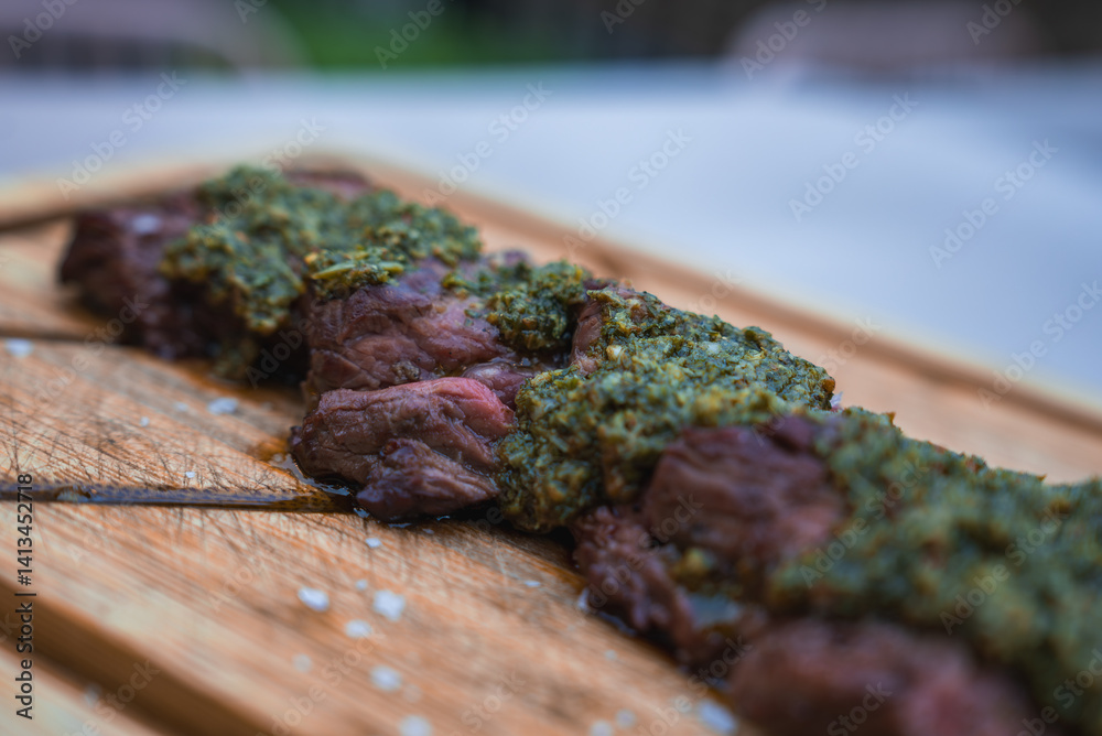 Slices of cooked meat topped with green herb based sauce on a wooden cutting board. The background shows blurred greenery, suggesting an outdoor setting.