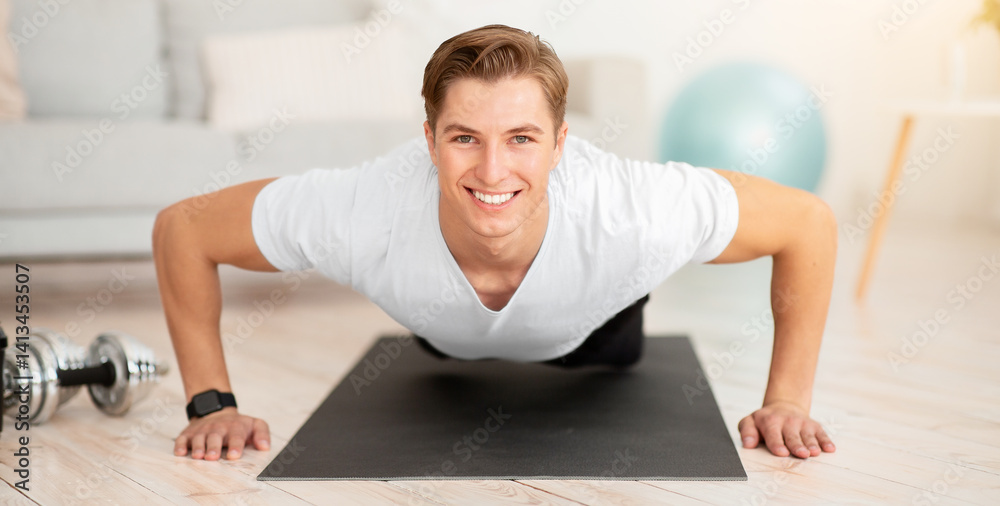 Fototapeta premium Healthy lifestyle at home during quarantine. Smiling handsome muscular guy doing push-ups on mat on floor in living room interior, free space