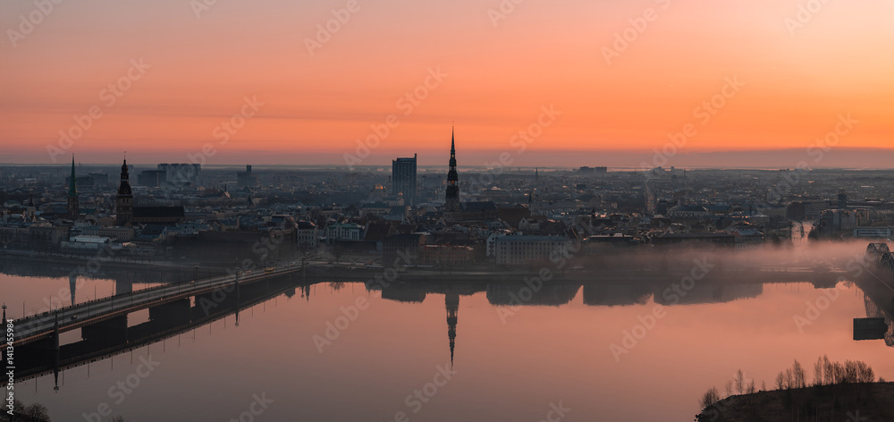 Serene sunrise over Riga, Latvia, featuring St. Peter's Church spire, historic buildings, a bridge over the Daugava River, and light morning mist.