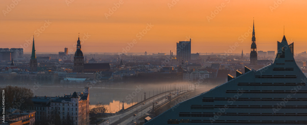 Serene sunrise over Riga, Latvia, featuring St. Peter's Church, Riga Cathedral, the National Library, and a misty Daugava River with a bridge.