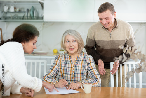 Thoughtful old woman compiling heritage document while middle-aged man and woman competing with each other in the kitchen