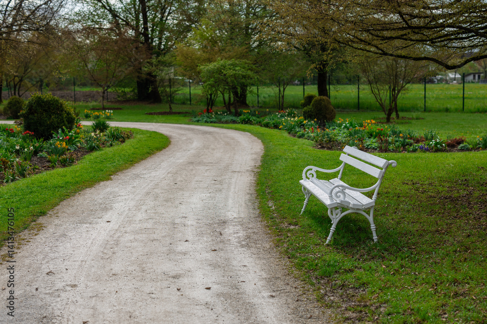 A serene park scene with a winding gravel path curving past a white ornate bench. The area is lush with green grass, colorful flower beds, and mature trees, creating a peaceful natural setting.