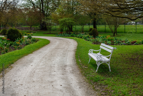 Obraz na płótnie A serene park scene with a winding gravel path curving past a white ornate bench. The area is lush with green grass, colorful flower beds, and mature trees, creating a peaceful natural setting. w salonie