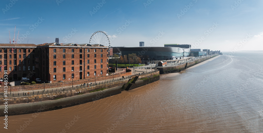 Aerial view of Liverpool showcasing Albert Dock's red brick warehouses, the Liverpool Wheel, MandS Bank Arena, and the River Mersey under a clear sky.