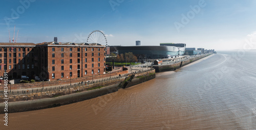Obraz na płótnie Aerial view of Liverpool showcasing Albert Dock's red brick warehouses, the Liverpool Wheel, MandS Bank Arena, and the River Mersey under a clear sky. w salonie