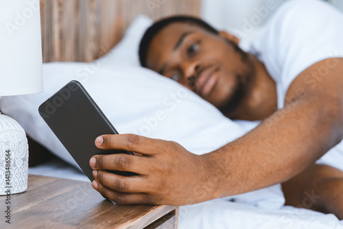 Black Man Turning Off Alarm Clock On Smartphone Waking Up Early In The Morning Lying In Bed At Home. Lack Of Sleep, Rest And Recreation Concept. Selective Focus, Shallow Depth