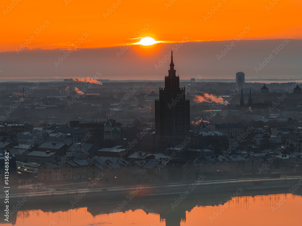 The Latvian Academy of Sciences spire stands silhouetted at sunrise in Riga, Latvia, with the Daugava River reflecting the warm orange sky.