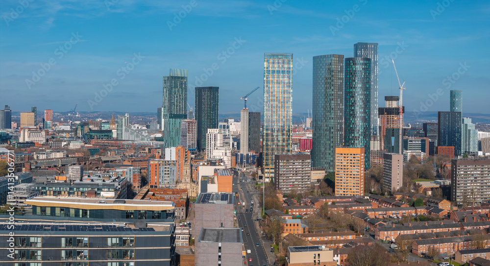 Aerial view of Manchester, UK, showing modern skyscrapers, construction cranes, and a mix of historic and contemporary buildings on a sunny day.
