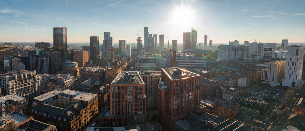 Aerial view of Manchester, UK, on a sunny day, featuring skyscrapers, construction cranes, historic brick buildings, and modern architecture.