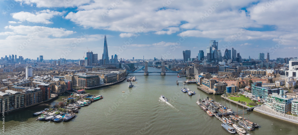 Aerial view of London featuring Tower Bridge over the River Thames, The Shard, Canary Wharf, and historic and modern riverside architecture on a bright day.