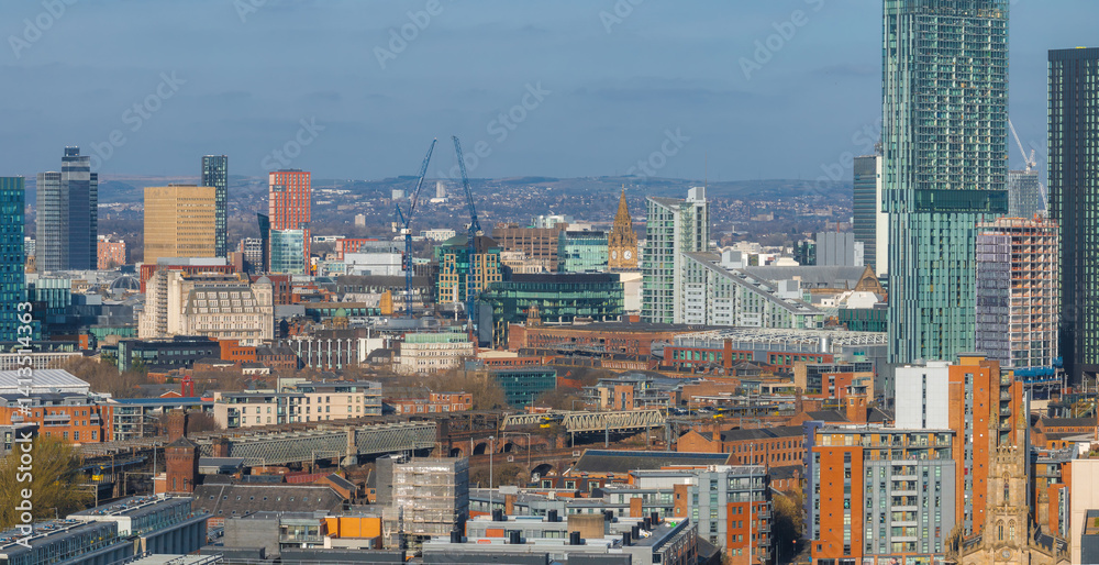 Aerial view of Manchester, UK, showcasing skyscrapers like Beetham Tower, the Town Hall clock tower, red brick buildings, and distant hills.