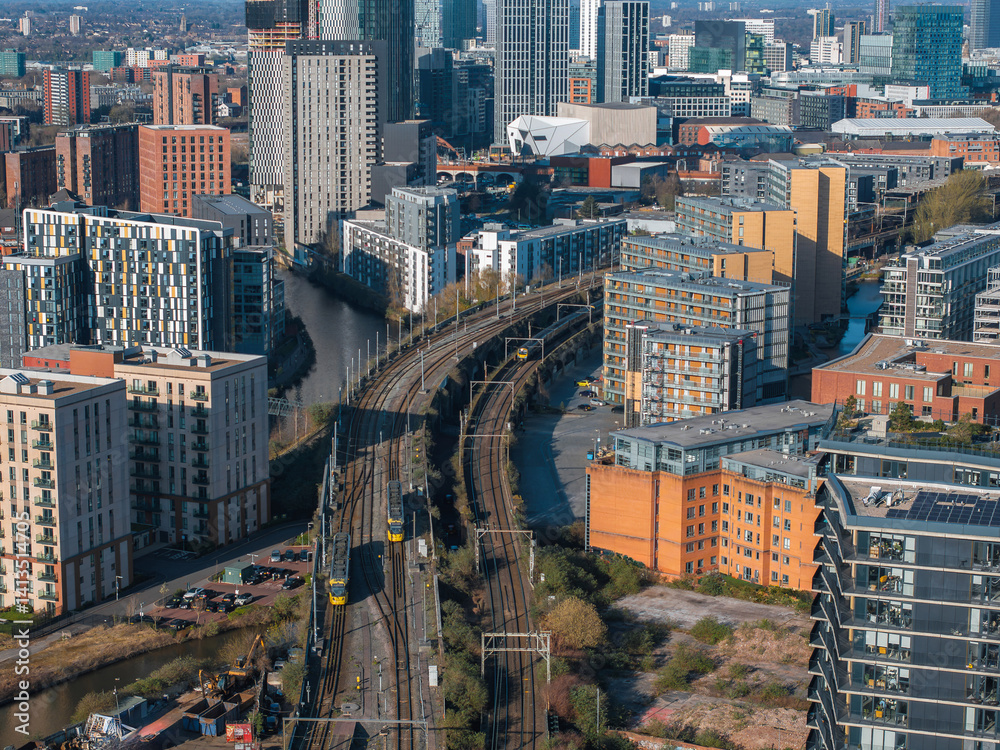 Aerial view of Manchester, UK, featuring modern skyscrapers, a tram line curving by a canal, and a mix of residential and commercial buildings.