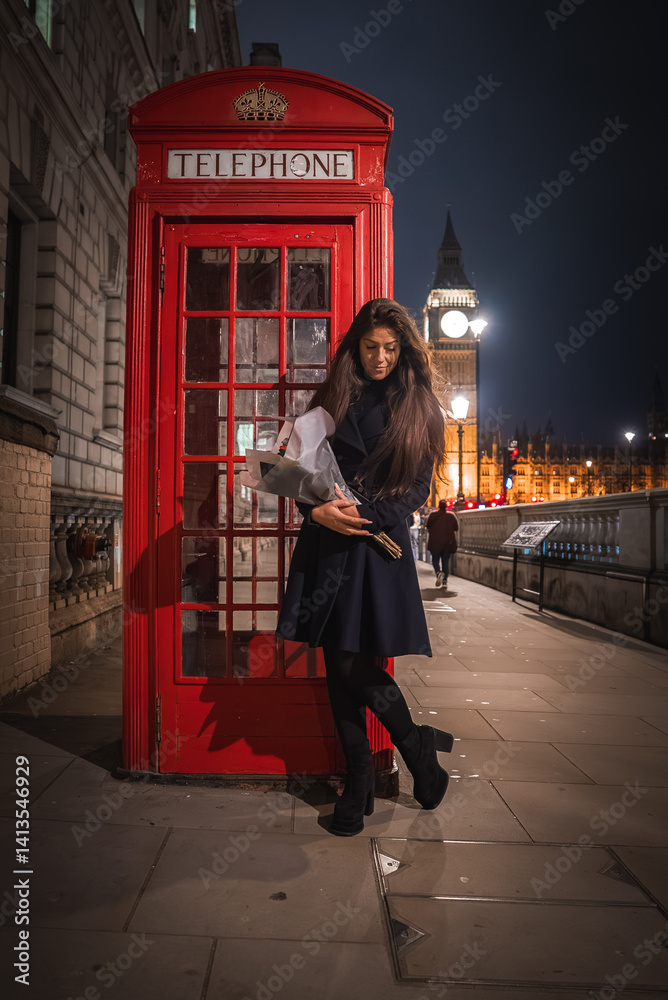 A red telephone booth stands in the foreground with a woman holding flowers. Big Ben and the Houses of Parliament are illuminated in the background.