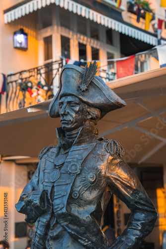 A bronze statue of a historical figure in a naval uniform with a tricorn hat stands outdoors. A building with balconies, flags, and awnings is in the background.