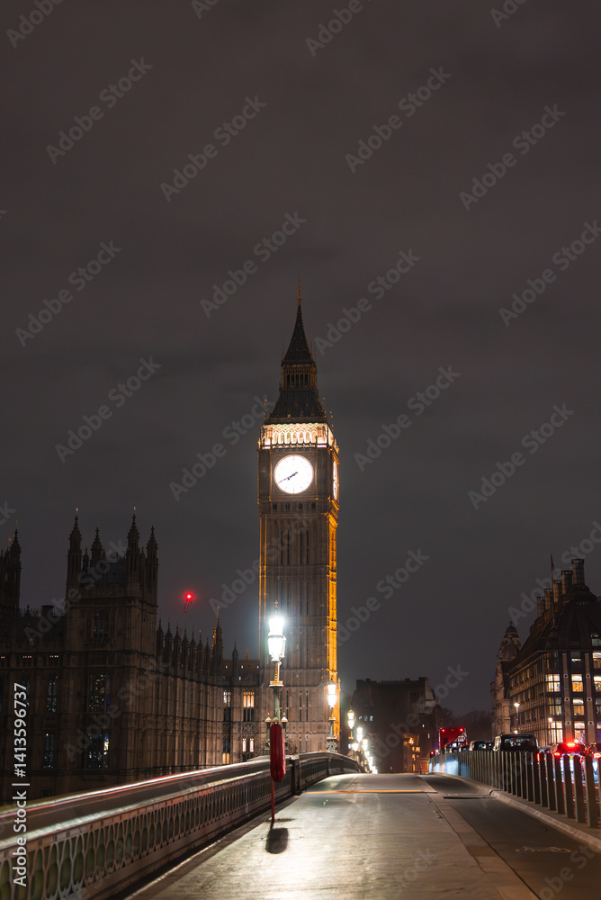 Obraz premium Big Ben's illuminated clock tower shines against a dark sky, with Westminster Bridge in the foreground featuring ornate railings and warm streetlights.