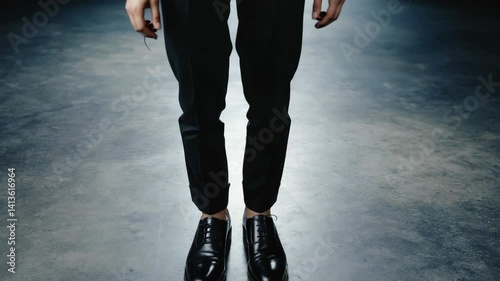 Stylish individual in dark trousers and shiny leather shoes standing on a minimalist, grey concrete floor in a studio setting.