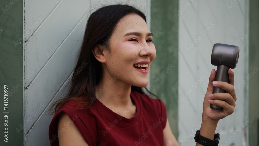 joyful asian woman laughing while holding a portable fan outdoors, dressed in red casual outfit with smartwatch, expressing freshness, self-care, beauty, energy and modern summer lifestyle joyful asian woman laughing while holding a portable fan outdoors, dressed in red casual outfit with smartwatch, expressing freshness, self-care, beauty, energy and modern summer lifestyle