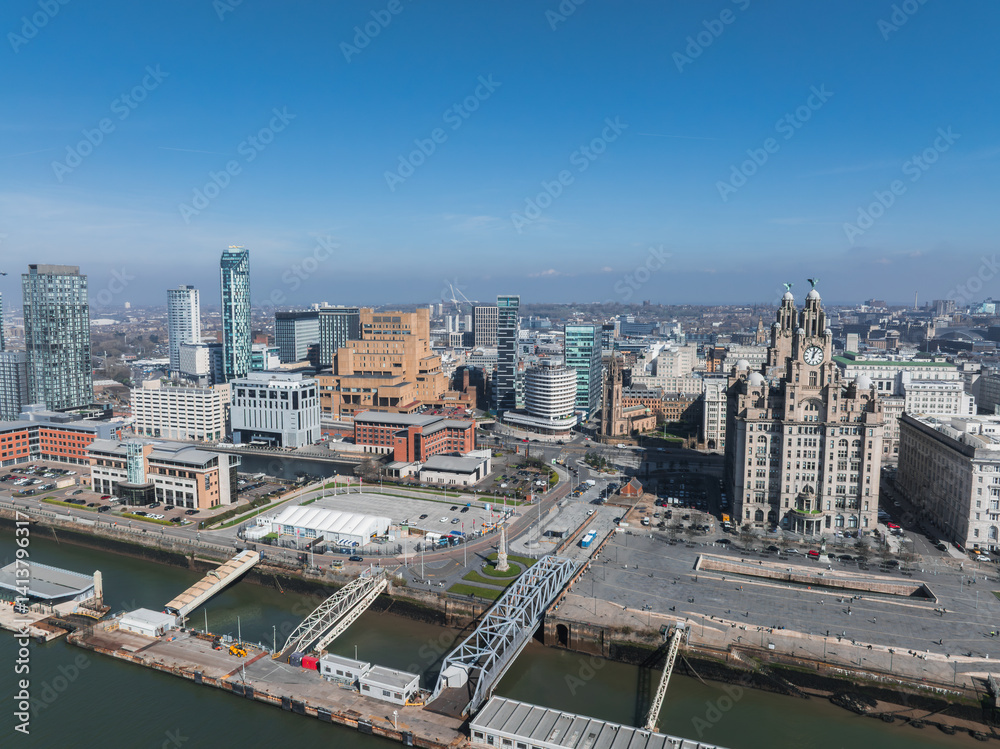 Aerial view of Liverpool featuring the Royal Liver Building, waterfront docks, a bridge, modern high rises, and historic architecture under a clear blue sky.
