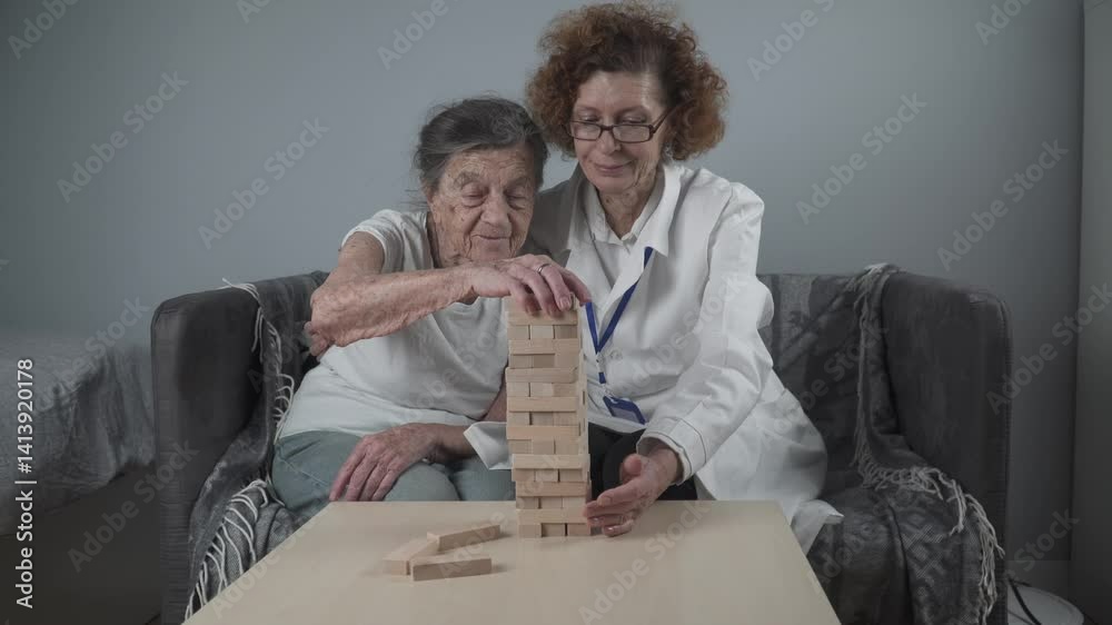 Jenga game. Theme is dementia, aging and games for old people. Caucasian senior woman builds tower of wooden blocks with the help of a doctor as part of a therapy and jenga game at a patient's home.