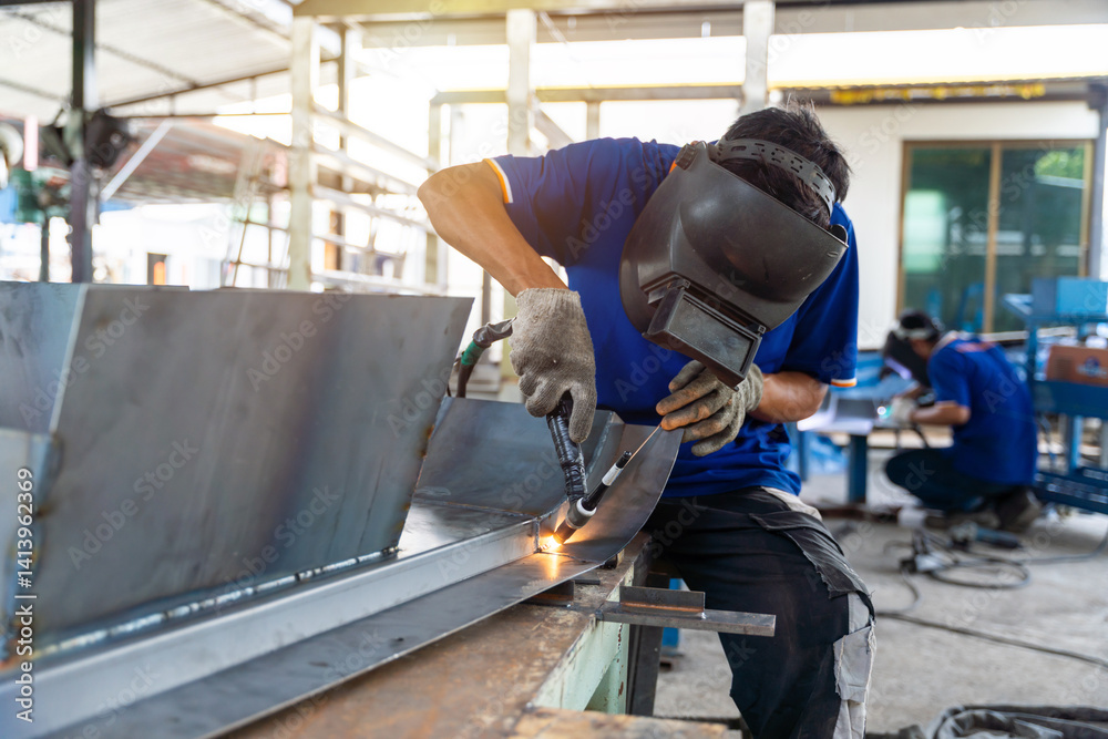 Skilled worker performing TIG welding on a curved metal structure in an industrial workshop, wearing safety gear. Another technician is working in the background. Skilled worker performing TIG welding on a curved metal structure in an industrial workshop, wearing safety gear. Another technician is working in the background.
