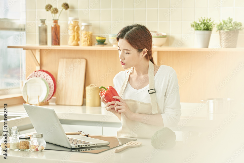 Young Woman Holding Bell Pepper and Using Laptop in Modern Kitchen Young Woman Holding Bell Pepper and Using Laptop in Modern Kitchen