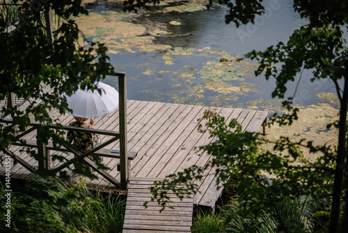 A person sits on a wooden dock beside a pond, holding a white umbrella, surrounded by trees and water lilies in a peaceful nature scene.. photo