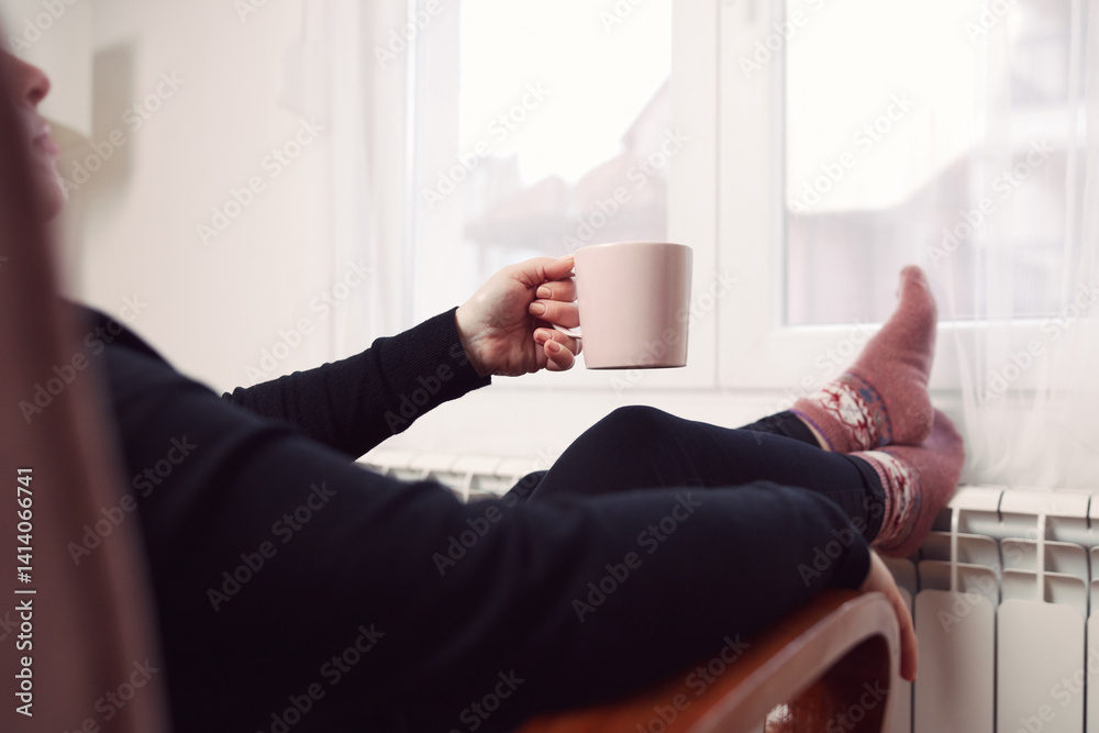 Woman drinking hot tea with her feet with socks on a home heating radiator on a cold winter day outdoors.