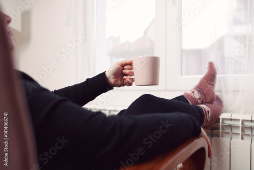 Woman drinking hot tea with her feet with socks on a home heating radiator on a cold winter day outdoors.