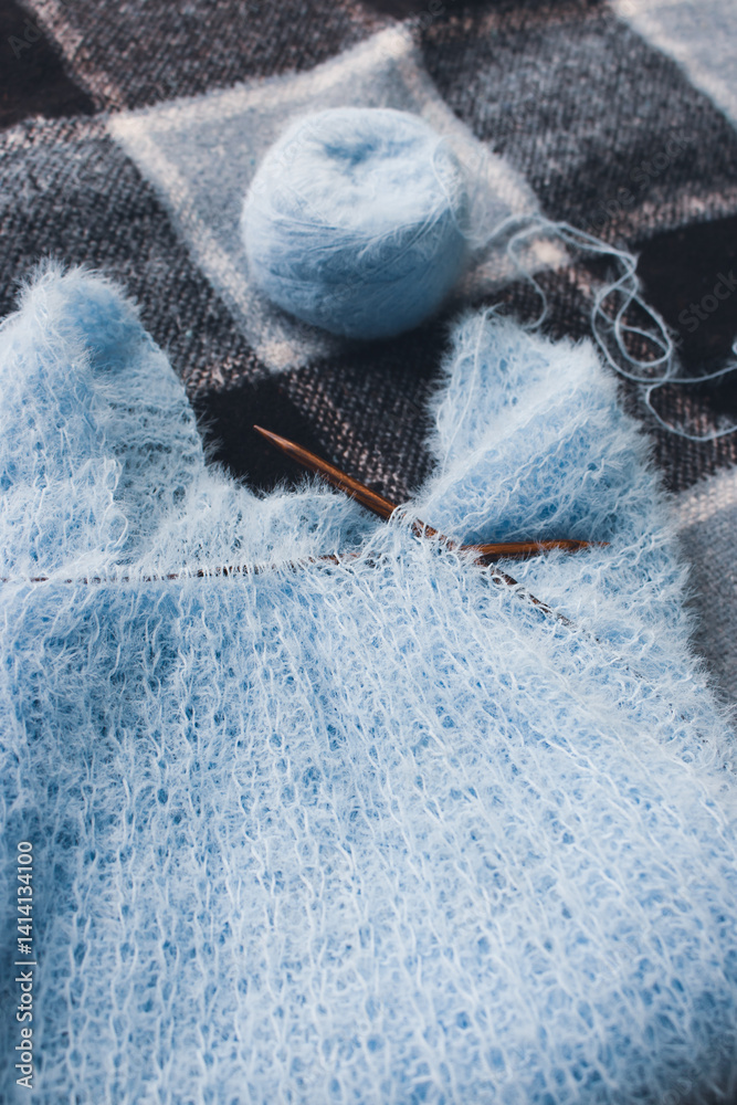 Blue cotton knitting with knitting needles on plaid. Soft blue knitwear with wooden circle needles. Knitting pattern background. Knitting lifestyle. Cozy hobby. Warm knitwear. Craft product concept