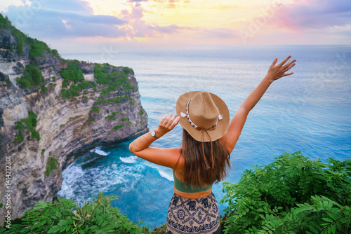 Traveler girl wearing hat enjoys turquoise stunning ocean view on cliff during traveling at beautiful destination on Bali island, Indonesia