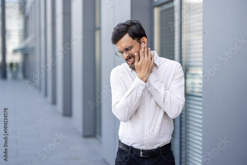 A man wearing glasses is experiencing pain while outdoors in a business environment, holding his face. He appears to be in discomfort.