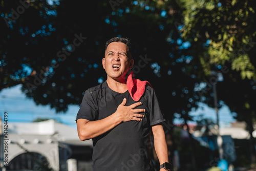 Southeast Asian man in his 50s clutches his chest in pain while walking outdoors, possibly experiencing angina or chest tightness from physical exertion.