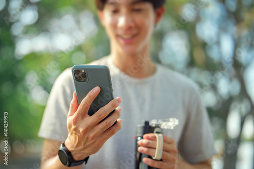 Close-up of male hand holding smartphone and water bottle at outdoor park – using fitness tracking app with wireless earbuds, healthy lifestyle and digital wellness