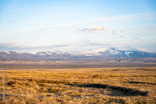 Obraz na płótnie Snow-capped mountain range under a soft blue sky, with golden fields stretching across a vast Icelandic landscape at sunset. w salonie
