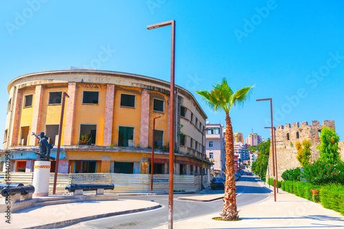 Street in Durres city from Albania with Former Hotel Volga. Sunlit street corner in Durres, Albania, featuring a curved, weathered building with boarded windows and a statue in the foreground