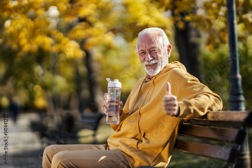 Portrait of senior sportsman sitting on a bench in park with bottle of water in hands and giving thumbs up at camera.