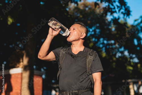 Middle aged Southeast Asian man hydrates with water from a bottle after finishing a challenging ruck in a sunny park.