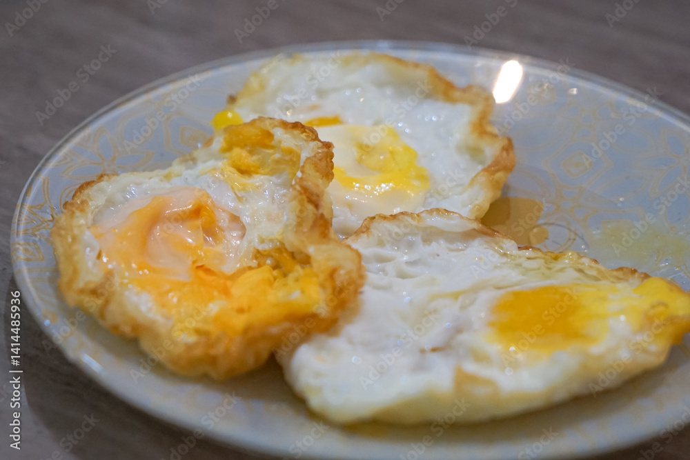 A plate with three fried eggs, featuring crispy edges and partially cooked yolks, arranged on a decorative floral-patterned plate placed on a wooden surface. A plate with three fried eggs, featuring crispy edges and partially cooked yolks, arranged on a decorative floral-patterned plate placed on a wooden surface.