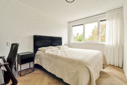 A sleek  modern bedroom featuring a bed with a plush white quilt  minimalist black furniture  large windows  and soft natural light streaming in.