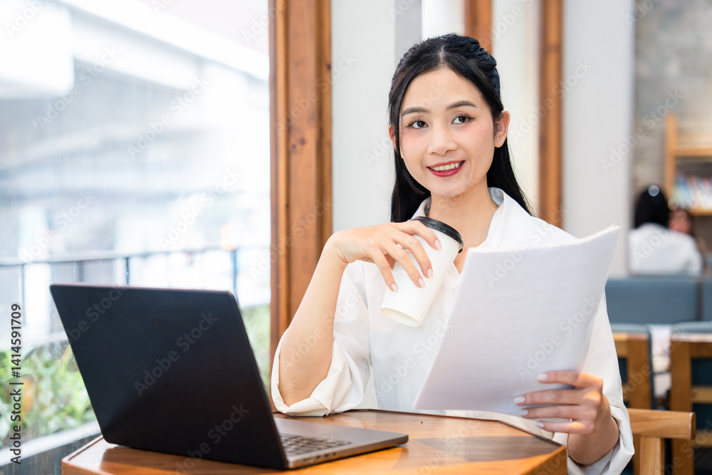 Smiling woman with laptop and paperwork enjoying a beverage at a cafe looking at camera Smiling woman with laptop and paperwork enjoying a beverage at a cafe looking at camera
