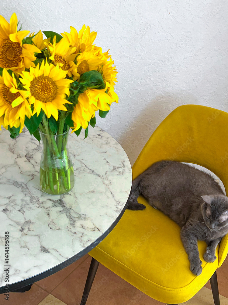 A grey cat is lying on a yellow armchair near a table with a bouquet of sunflowers