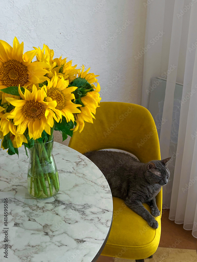 A grey cat is lying on a yellow armchair near a table with a bouquet of sunflowers